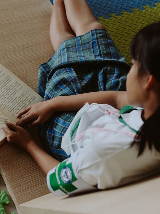 A young girl in a school uniform reading a book while sitting on a colorful mat indoors.